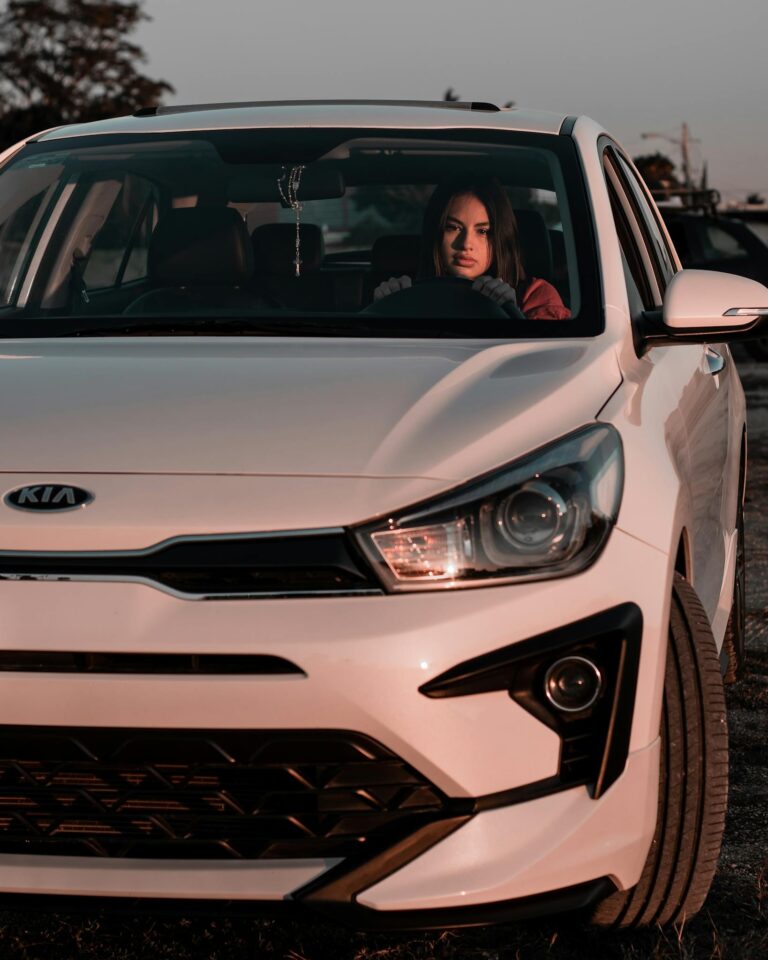 A woman sitting in a white Kia car, captured in warm evening light outdoors.